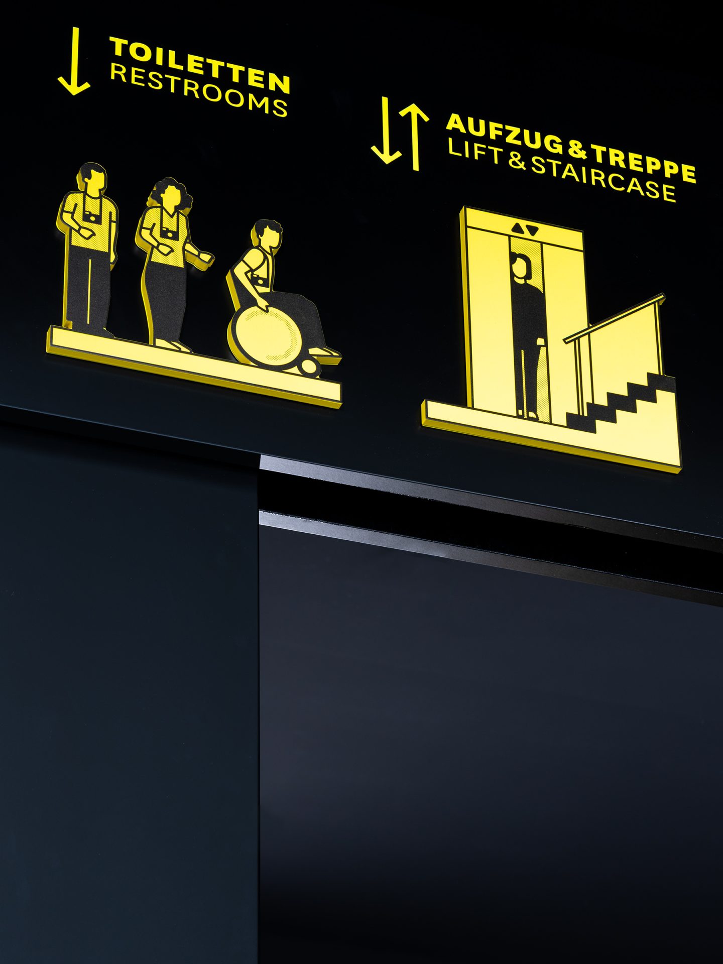 Ceiling-mounted wayfinding sign for Haus des Tourismus Stuttgart, featuring bilingual restroom, lift and staircase directions with custom yellow pictograms on a dark interior background.
