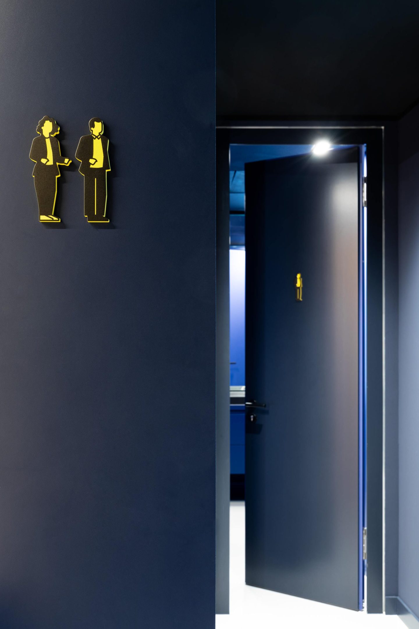 Restroom pictogram for Haus des Tourismus Stuttgart, featuring a custom yellow visitor icon applied to a dark blue door as part of the interior wayfinding system.