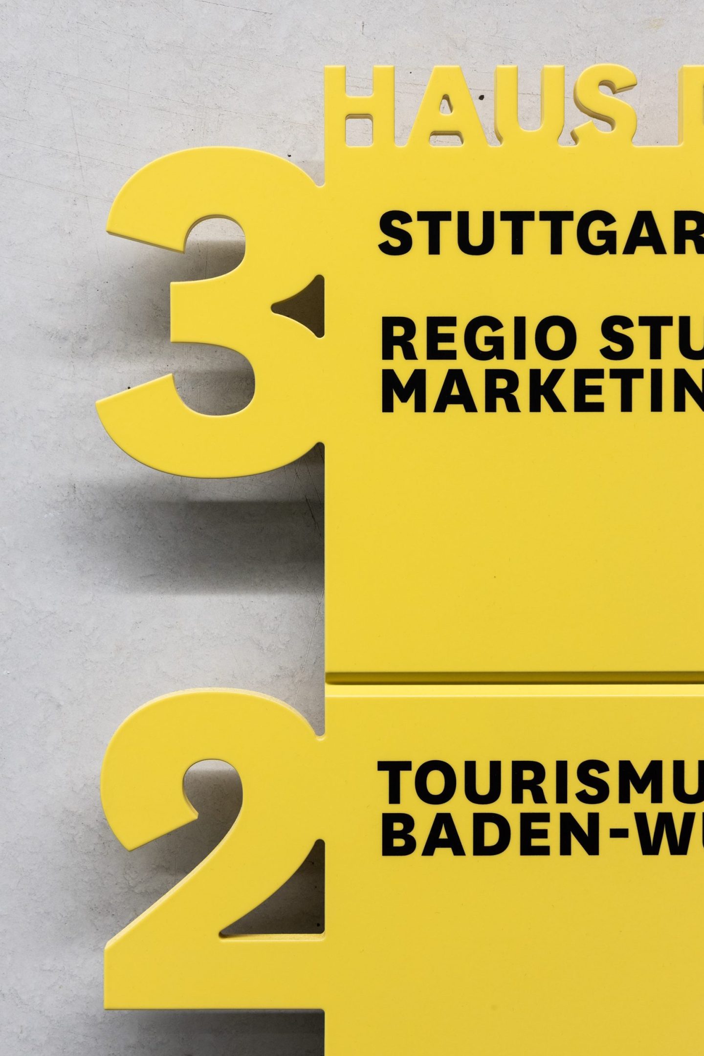 Stairwell wayfinding sign for Haus des Tourismus Stuttgart, featuring bold yellow floor markers with large cut-out numbers and black destination typography as part of the interior orientation system.